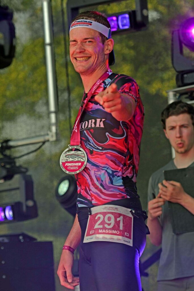 Finisher Apfelland Triathlon Massimo Köstl-Lenz als Triathlet und Ausdauer Coach nach der geschafften Mitteldistanz beim Apfelland Triathlon am Stubenbergsee, strahlend mit Finisher-Medaille auf der Bühne und Sinnbild für sein Sport- und Ernährungscoaching.