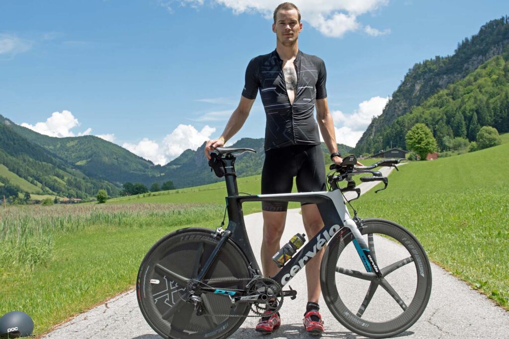 Triathlon Coach Massimo Köstl-Lenz steht mit seinem Zeitfahrrad vor der Bergkulisse in Tirol und besichtigt am Vortag des Triathlons die Strecke.