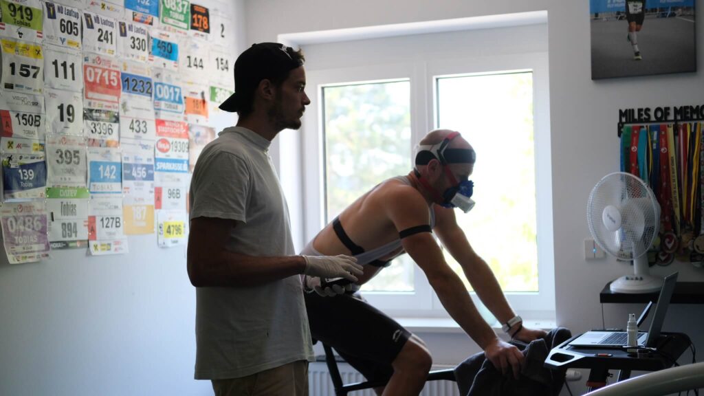 Sportwissenschaftler Massimo Köstl-Lenz begleitet einen Athleten mit Atemmaske beim VO₂max-Leistungstest am eigenen Fahrrad und setzt präzise Leistungsdiagnostik im Ausdauersport ein.