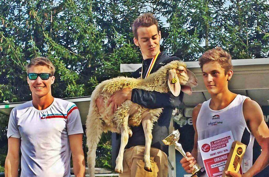 Massimo Köstl-Lenz gewinnt den Triathlon in Kraig und erhält als Preis ein Schaf, das er bei der Siegerehrung im Arm hält.