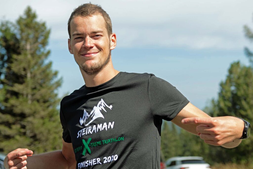 Massimo Köstl-Lenz trägt das Steiraman Styrian Xtreme Triathlon Finisher T-Shirt und zeigt lachend auf die Aufschrift.