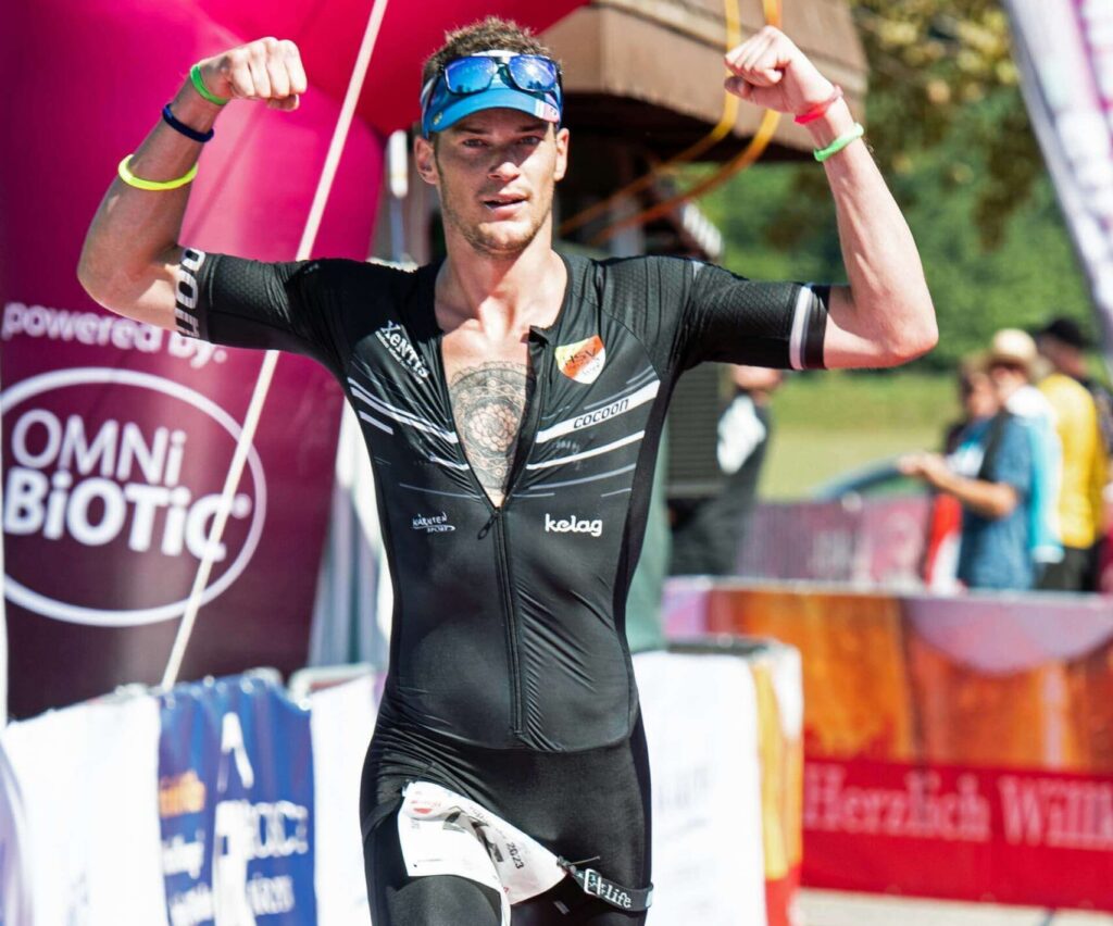 Massimo Köstl-Lenz läuft beim Südkärntner Triathlon am Klopeiner See über die Halbdistanz mit erhobenen Armen ins Ziel und zeigt sich als aktiver Leistungssportler.