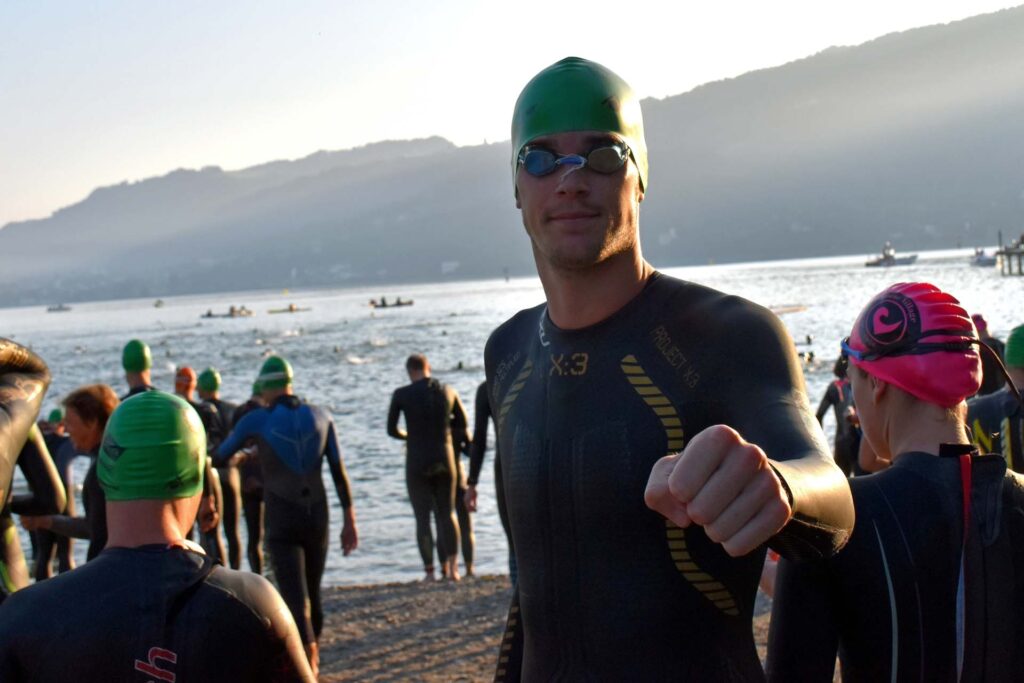 Massimo Köstl-Lenz steht vor dem Start beim Triathlon Trans Vorarlberg im Neoprenanzug am See, umgeben von anderen Athleten im Morgenlicht.