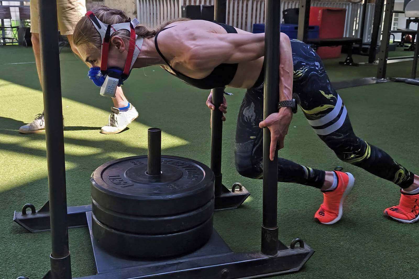 Hyrox-Sportlerin beim Sled Push mit VO2-Master-Maske während einer mobilen Leistungsdiagnostik direkt in der Wettkampf-Simulation im Gym.