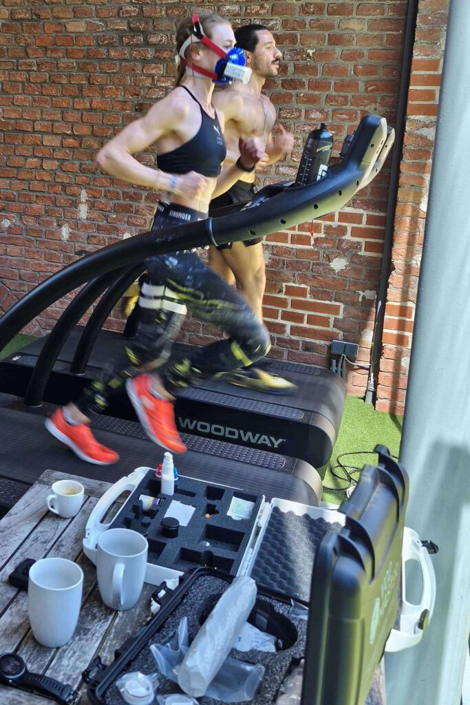 Hyrox-Sportlerin läuft im Gym mit VO2-Master-Maske am Laufband, während das mobile Test- Equipment der Leistungsdiagnostik im Vordergrund steht.