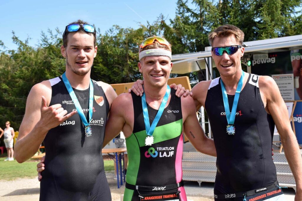 Triathlon Trainer Massimo Köstl-Lenz gehört beim Gösselsdorfer Triathlon in Kärnten zu den drei schnellsten Athleten des Tages; auf dem Foto sind die drei bei der Siegerehrung mit Medaillen zu sehen.